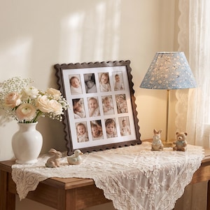 May include: A dark brown scalloped picture frame holding twelve individual baby portraits, displayed on a table with a white vase of pale pink roses, a blue floral lamp, and two small ceramic animal figurines on a lace tablecloth.