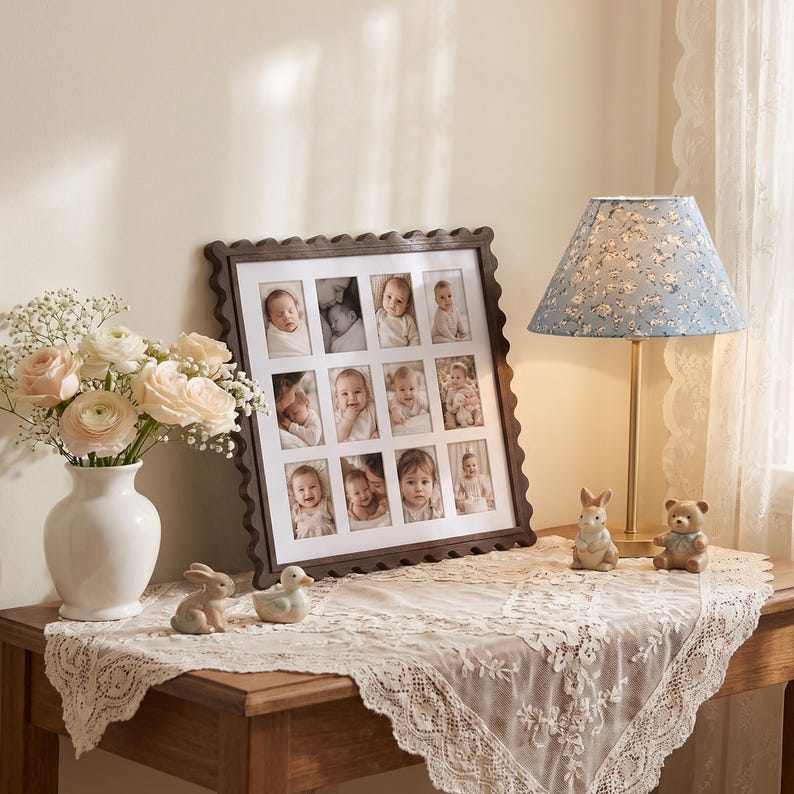 May include: A dark brown scalloped picture frame holding twelve individual baby portraits, displayed on a table with a white vase of pale pink roses, a blue floral lamp, and two small ceramic animal figurines on a lace tablecloth.