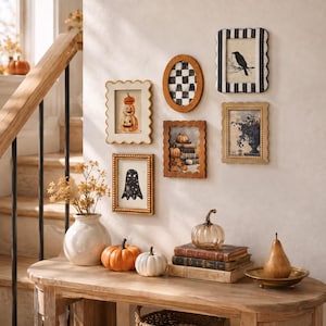 Op de afbeelding: Een galerijwand met vijf decoratieve lijsten met seizoensgebonden kunst, waaronder een geest, pompoenen en een kraai. Op de houten sidetable staan een witte vaas met droogbloemen, twee pompoenen, een glazen pompoen, boeken en een peer.