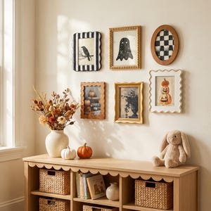 Könnte beinhalten: Ein Holzkonsolentisch mit gewellter Kante und einer Bilderwand darüber mit sechs gerahmten Drucken, darunter eine Krähe, ein Geist, ein Schachbrettmuster, Kürbisse und Laub. Eine Vase mit Trockenblumen, zwei kleine Kürbisse und ein Plüschhase.