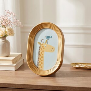 K&ouml;nnte beinhalten: Ein ovaler goldener Bilderrahmen mit gerippter Textur. Der Rahmen enth&auml;lt eine Illustration einer gelben Giraffe mit braunen Flecken und einem kleinen blauen Vogel, der auf ihrem Kopf sitzt, vor einem hellblauen Hintergrund.