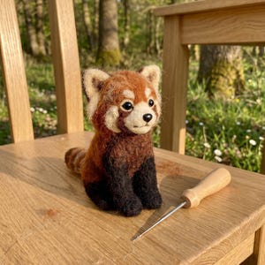 May include: A handmade felt red panda figure with a detailed design. The red panda has a brown and black body, a white face, and a striped tail. A wooden-handled tool rests on the wooden chair next to the figurine.