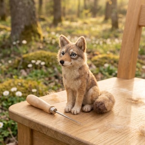 May include: A needle-felted fox figurine with brown and cream fur, dark eyes, and a pointed nose, sitting upright. Beside the fox is a wooden-handled felting needle with a thin metal tip, both resting on a light-colored wooden surface.
