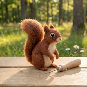 Könnte beinhalten: Ein handgefertigtes Eichhörnchen aus Filz, hauptsächlich braun mit cremefarbenem Bauch, auf einer Holzoberfläche. Ein Nadelwerkzeug mit Holzgriff liegt daneben. Der Hintergrund zeigt eine verschwommene Waldszene.