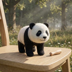 Könnte beinhalten: Handgefertigte Panda-Bärenfigur aus Filz mit schwarzen und weißen Details, steht auf einem Holzstuhl. Der Panda hat schwarze Ohren, Augen und Gliedmaßen. Ein Holz-Nadelwerkzeug liegt auf dem Stuhl.