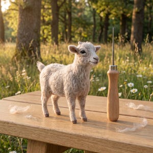 Op de afbeelding: Een naaldgevilte lichtbeige geitenfiguur met een textuur van wol, staand naast een metalen viltgereedschap met een houten handvat. De geit heeft kleine oren, donkere ogen en een korte staart, met losse plukjes wol verspreid op het oppervlak.