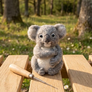 May include: A handmade gray koala bear figurine with a white belly sits on a wooden bench. A wooden-handled awl lies next to the koala. The background is a blurred forest scene with sunlight.