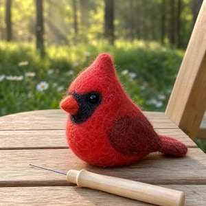 May include: A handcrafted, red felt cardinal bird with black accents and a wooden needle tool. The bird has a rounded body, a small beak, and a detailed wing. The needle tool is light-colored wood. The scene is set outdoors with a blurred background of trees and sunlight.