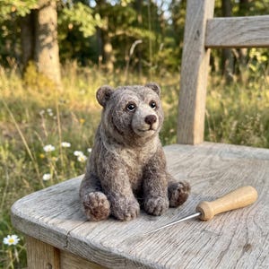 May include: A needle-felted brown bear figurine with a textured, fuzzy coat, dark eyes, and a small nose, sitting upright. Beside the bear lies a metal felting needle with a light-coloured wooden handle, resting on a wooden surface.