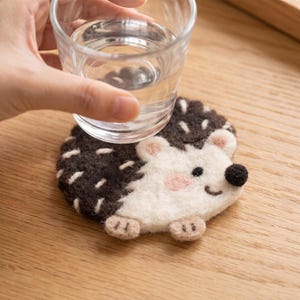 May include: A round felt coaster shaped like a hedgehog with a dark brown back featuring white stitch details, a cream-coloured face, a small black nose, pink cheeks, and two small beige feet. A clear glass of water rests on top of the coaster.