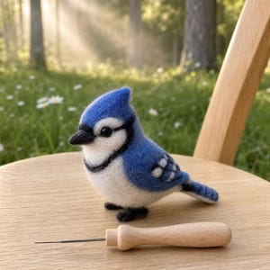 Peut inclure: Figurine artisanale en feutre représentant un geai bleu, avec des détails blancs, un bec et des pattes noirs, et un outil à aiguille en bois. L'oiseau est posé sur une chaise en bois.