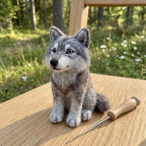 May include: A handcrafted gray and white felt wolf figurine with a detailed design. The wolf is sitting on a wooden surface. A wooden-handled needle tool is next to the wolf. The background includes trees and greenery.