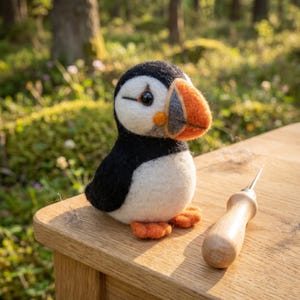 Puede incluir: Un frailecillo de fieltro hecho a mano con una combinación de colores negro, blanco, naranja y gris. El ave está posada sobre una superficie de madera junto a una herramienta de madera. El fondo es una escena borrosa del bosque.