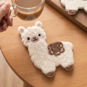 Puede incluir: Un posavasos de lana afieltrada de color crema con forma de alpaca. El posavasos presenta una mancha marrón en el lomo, pezuñas marrones, ojos negros, nariz negra y mejillas rosadas. La textura es difusa y suave con un contorno definido.