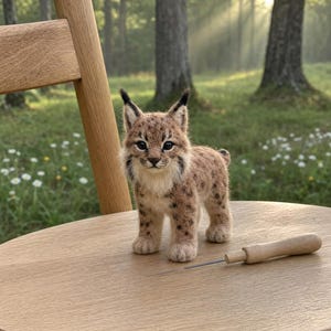 May include: A handmade felted lynx figure with detailed markings and black ear tips sits on a wooden chair. A needle tool is placed close by. The background shows a blurred forest scene with sunlight.