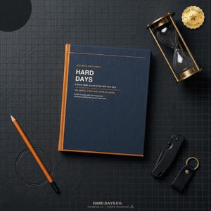 Puede incluir: Un diario azul marino con el texto "HARD DAYS" y "A Mental Health Journal for Men Who Work Hard." Un reloj de arena, un lápiz, un cuchillo y un llavero están dispuestos sobre una superficie de rejilla negra. Una insignia de "Top Seller" es visible.