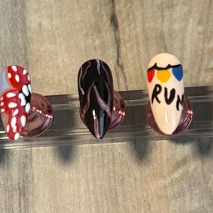 May include: Five artificial nails with unique designs. One nail is white with the number 11, another has a red and white pattern, a third is black, the fourth has the word "RUN", and the last is blue with "SCOOP AHO".