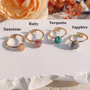 Puede incluir: Cuatro anillos dorados con banda abierta, cada uno con una piedra preciosa en bruto sin tallar. Las piedras están etiquetadas como Sunstone, Ruby, Turquoise y Sapphire. Cada anillo muestra un mineral de forma natural sobre una banda fina.