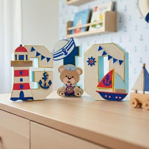 Puede incluir: Artículos decorativos de madera con temática náutica, que incluyen un faro, los números "30", un oso de peluche y un velero. Los artículos están pintados en rojo, blanco, azul y dorado. El fondo presenta un papel pintado náutico y estanterías de madera.
