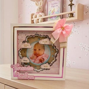Può includere: Una cornice bianca per foto di neonati con un fiocco rosa. La cornice ha un bordo smerlato attorno alla foto del bambino, con dettagli sulla data di nascita, l'ora, il peso e l'altezza del bambino. Il nome "Florencia Isidora" è scritto sulla cornice.