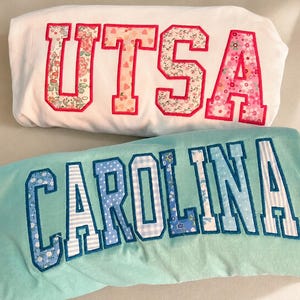 May include: Two folded t-shirts. The top white shirt features the letters UTSA in pink-bordered floral fabric. The bottom light blue shirt features the letters CAROLINA in blue-bordered fabric with various blue and white patterns including stripes.