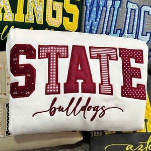 May include: A collection of five folded sweatshirts featuring large collegiate-style block letters with patterned fabric appliques. The sweatshirts are white, dark green, heather gray, tan, and charcoal, displaying text including VIKINGS, WILDCATS, STATE bulldogs, and aztecs.