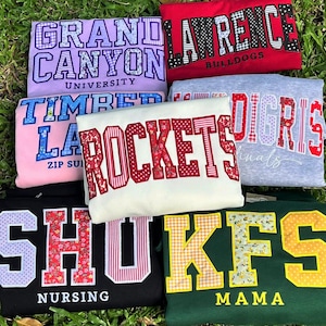 May include: A collection of seven folded sweatshirts in various colors, each featuring large collegiate-style block letters with diverse fabric patterns. The shirts display text including Grand Canyon University, Lawrence Bulldogs, Timber Lake, Rockets, Digris Cardinals, SHU Nursing, and KFS Mama.