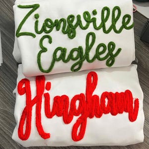 May include: Two white sweatshirts featuring textured chenille lettering. The top sweatshirt displays the green text Zionsville Eagles in a cursive font. The bottom sweatshirt features the red text Hingham in a matching cursive script style.