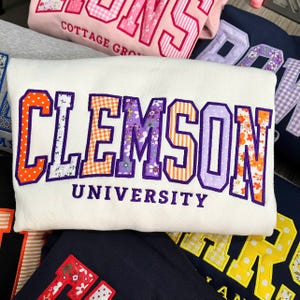 May include: A white sweatshirt featuring the word CLEMSON in large, block-style letters with a purple outline. Each letter is filled with a different patterned fabric, including orange polka dots, floral prints, and orange and white gingham checks.