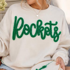 May include: A cream-colored crewneck sweatshirt featuring the word Rockets embroidered in a textured, fuzzy green yarn across the chest. The design is executed in a cursive script style with a thick, raised appearance on the fabric surface.