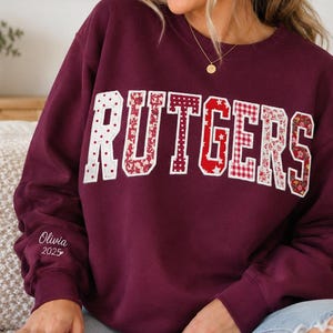 May include: A maroon long-sleeve sweatshirt featuring the word RUTGERS in large, block-style letters. Each letter has a unique white and red patterned fill, including polka dots, florals, and gingham. The name Olivia 2025 is embroidered on the sleeve.