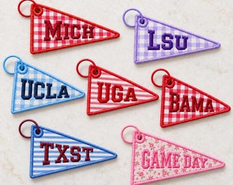 May include: Seven triangular embroidered keychains with metal rings. Designs include red gingham with MICH, purple gingham with LSU, light blue gingham with UCLA, red striped with UGA, red gingham with BAMA, blue striped with TXST, and pink floral with GAME DAY.