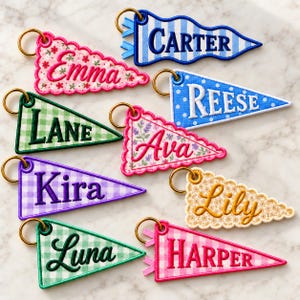 May include: A collection of nine triangular embroidered pennant keychains featuring various patterns and colors. Each keychain displays a unique name: Carter, Emma, Reese, Lane, Ava, Kira, Lily, Luna, and Harper, attached to a gold-toned metal ring.