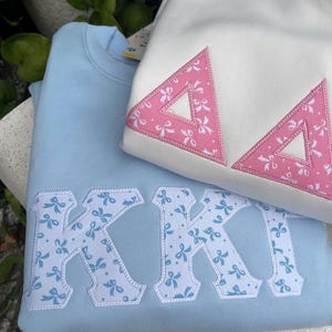 May include: A light blue sweatshirt featuring large white letters with a blue bow pattern and a white sweatshirt featuring two pink triangular patches with a white bow pattern. Both garments have stitched edges around the letters and patches.