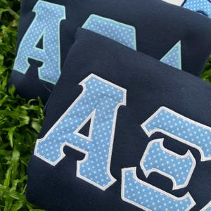 May include: A navy blue sweatshirt featuring large, block-style Greek letters appliqued on the front. The letters are light blue with a white polka dot pattern, outlined in a thin light blue border and a thicker white stitched edge.