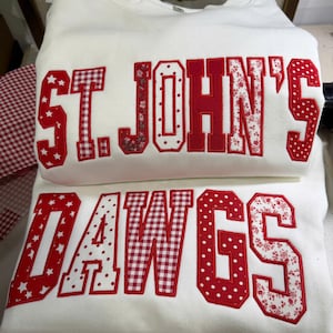 May include: A white crewneck sweatshirt featuring large, red, fabric-appliqued letters that spell out ST. JOHN'S DAWGS. The letters are decorated with various patterns including red and white gingham, polka dots, stars, and floral prints.