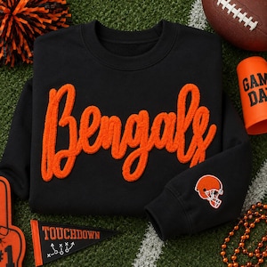 May include: A black crewneck sweatshirt featuring the word Bengals in large, textured orange chenille script lettering across the chest and a small orange and white football helmet logo embroidered on the right sleeve cuff.
