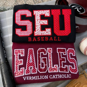 May include: A black sweatshirt with red applique letters reading SEU BASEBALL and a maroon sweatshirt with white applique letters reading EAGLES VERMILION CATHOLIC. The letters feature various red and white patterns including gingham, floral, and polka dots.