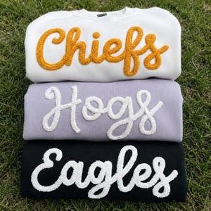 May include: Three crewneck sweatshirts stacked vertically. The top is white with gold chenille lettering reading Chiefs. The middle is light purple with white chenille lettering reading Hogs. The bottom is black with white chenille lettering reading Eagles.