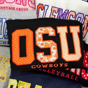 May include: A black sweatshirt featuring the letters OSU in large, fabric-appliqued block font. The O has orange polka dots, the S has an orange floral pattern, and the U has orange stripes. The word COWBOYS is embroidered in orange below the letters.