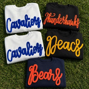 May include: Five crewneck sweatshirts featuring raised, textured lettering. Two white sweatshirts display blue 'Cavaliers' text. One black sweatshirt features orange 'Thunderhawks' text, one black sweatshirt has yellow 'Deacs' text, and one navy sweatshirt has orange 'Bears' text.