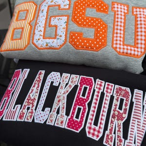 May include: A grey sweatshirt featuring the orange and white patterned applique letters BGSU, stacked above a black sweatshirt with the pink and white patterned applique letters BLACKBURN, both featuring varsity-style block lettering.