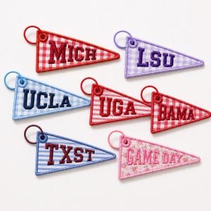 May include: Six triangular pennant keychains featuring embroidered text including MICH, LSU, UCLA, UGA, BAMA, TXST, and GAME DAY. Each keychain has a coordinating colored metal ring and displays various gingham, striped, or floral fabric patterns.