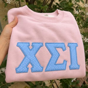 May include: A light pink crewneck sweatshirt featuring large, appliqued Greek letters in a light blue fabric with a white polka dot pattern. The letters are outlined with a matching light blue stitched border, creating a textured, layered look.