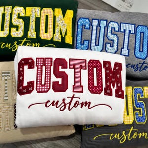 May include: A collection of five folded sweatshirts in white, dark green, heather gray, tan, and charcoal gray. Each sweatshirt features the word CUSTOM in large block letters with patterned fabric appliques and the word custom in script.