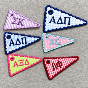 May include: Six triangular embroidered patches with scalloped edges and eyelets. The patches feature various patterns and Greek letters: purple stripes with ΣΚ, blue gingham with ΑΔΠ, blue polka dots with ΑΔΠ, pink polka dots with ΧΩ, lime green polka dots with ΑΞΔ, and red gingham with ΑΦ.