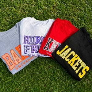 May include: Four t-shirts in gray, white, red, and black. The gray shirt features orange text reading BARK UNIVERSITY. The white shirt has purple text reading HORN FROGS Give 'Em Hell. The red shirt has patterned text. The black shirt has yellow text reading JACKETS.