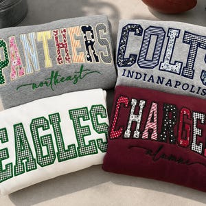 May include: Four folded sweatshirts featuring collegiate-style applique lettering. One gray sweatshirt reads PANTHERS northeast, one gray reads COLTS INDIANAPOLIS, one white reads EAGLES, and one maroon reads CHARGERS alumni, all with patterned fabric letters.