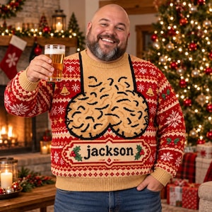 Könnte beinhalten: Ein beigefarbener und roter langärmeliger Strickpullover mit einem festlichen Weihnachtsmuster aus Schneeflocken, Stechpalmen und Glocken. In der Mitte des Pullovers befindet sich ein großes Grafikdesign mit dem Namen jackson in schwarzen Kleinbuchstaben.