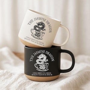 Puede incluir: Una pila de dos tazas de cerámica, una crema y otra negra, cada una con un gráfico de un dragón saliendo de una taza de té con el texto The Jasmine Dragon, Uncle Iroh's Tea House, Quality Teas of Ba Sing Se impreso en la parte frontal.